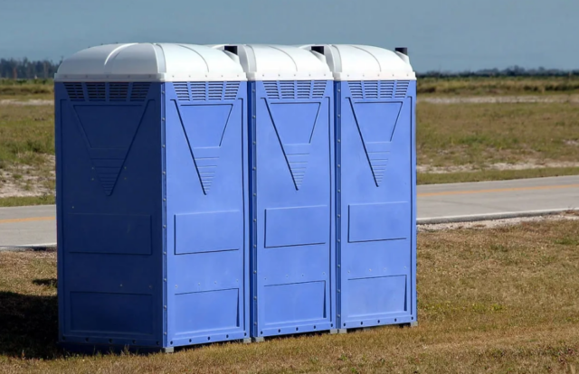 Three blue portable toilets lined up on a grassy roadside area, ready for use at outdoor events, construction sites, or remote locations.