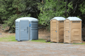 ADA-compliant gray portable toilet alongside two standard tan porta potties in a wooded outdoor setting, suitable for weddings, events, or campground use