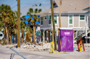 Purple portable toilet stationed in a hurricane-damaged coastal area with debris and raised homes, providing essential sanitation during disaster relief efforts